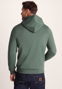 Man wearing a plain green hoodie and dark blue jeans facing away against a light beige background.