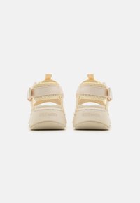 Beige sneakers with a textured upper, adjustable straps, and a sturdy rubber sole. Features branding on the heel and a pull tab.