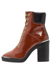 Bottine en cuir verni marron avec un bout pointu, œillets dorés, accents noirs sur le talon, et un talon épais noir.