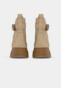Beige semskede ankelstøvler med justerbare remmer og en brun gummisål, med en afrundet tå og en let hævet hæl.