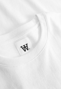 Fehér pamut póló kerek nyakkivágással és bordázott gallérral; a gallér alján fekete címke található, rajta a "W" betűvel.