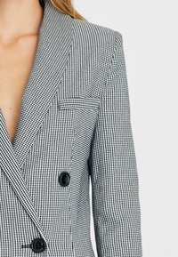 Veste croisée noir et blanc à motif pied-de-poule, avec un col à revers cranté, deux boutons noirs et une poche poitrine.