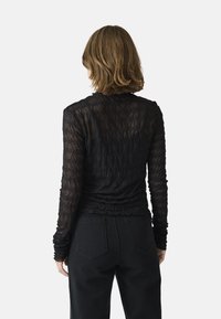 Svart långärmad topp gjord av genomskinlig texturerad tyg med ett mönstrat design. Tätt sittande siluett med rynkade detaljer vid midjan och ärmarna.