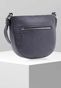 Czarna skórzana torba na ramię o teksturowanej powierzchni z przednią kieszenią na zamek błyskawiczny, regulowanym paskiem oraz dyskretnym wytłoczonym logo, umieszczona na białym piedestale z neutralnym tłem.