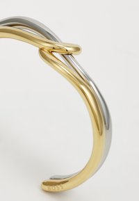 Pulsera de muñeca de metal que presenta bandas entrelazadas en oro y plata, superficie lisa, diseño minimalista y nombre de la marca "BOSS" grabado en el extremo.