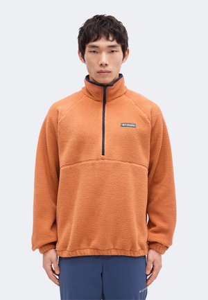 Jeune homme portant un pull polaire orange à fermeture éclair partielle avec logo Columbia, associé à un pantalon bleu sur un fond clair uni.
