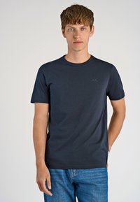 Mörkblå kortärmad t-shirt i mjukt tyg, med rund halsringning och en subtil logotyp på bröstet. Bärs med blå jeans.