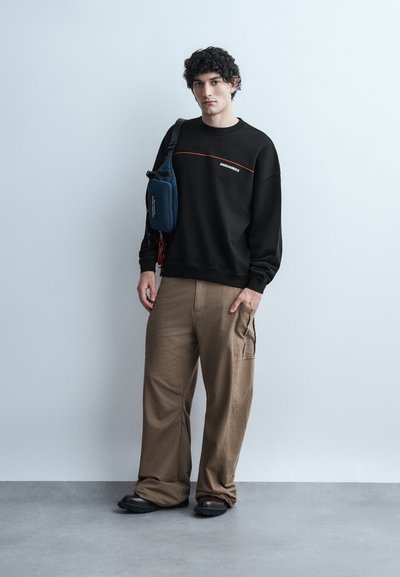 Άνδρας με σγουρά μαλλιά φορά μαύρο φούτερ, χακί άνετο παντελόνι cargo, μαύρα παπούτσια και μπλε σκούρο τσάντα χιαστί, στέκεται μπροστά σε απλό ανοιχτό γκρι τοίχο.