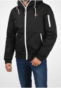 Veste bomber noire avec fermeture éclair blanche, capuche ajustable, poches latérales et poignets côtelés. Confectionnée en tissu lisse et résistant avec un accent logo.