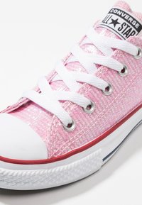 Różowe płócienne trampki o teksturowanym, połyskującym wykończeniu, z białymi sznurówkami, gumowym noskiem i czerwonym akcentem wzdłuż podeszwy. Posiadają metalowe oczka.