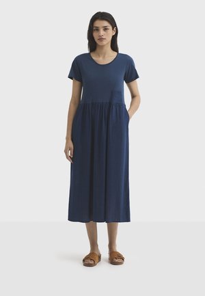 Jonge vrouw staat frontaal en draagt een marineblauwe midi-jurk met korte mouwen, een geplooide rok en bruine slippers.