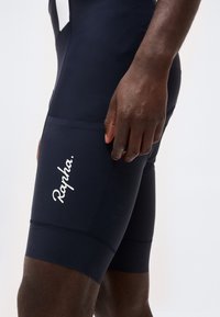 Κοντινό πλάνο ενός ατόμου που φοράει μαύρα ποδηλατικά παντελόνια Rapha, εστιάζοντας στην περιοχή του μηρού και το χέρι που ακουμπά δίπλα.