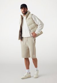 Beige padded vest with hood over a white hoodie, paired with beige shorts featuring a logo patch and cream sneakers. Simple, casual design.