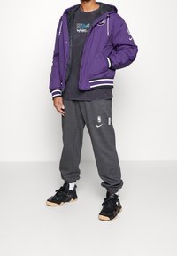 Lila huva jacka med vitrandiga ärmar, grå grafisk t-shirt, grå sweatpants med logotyp, och svarta sneakers med gummisula.