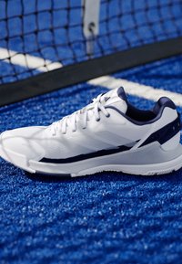 Bílé atletické boty s texturovaným svrškem, námořnickými akcenty a aerodynamickým designem. Uloženy na syntetické modré trávě blízko sítě.