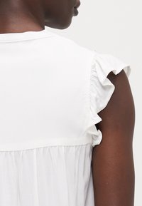 Blusa bianca con maniche corte a balze, schiena arricciata e una texture liscia. Design semplice e pulito, senza motivi visibili.