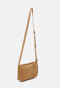Sandfärgad mocka crossbody-väska med en lång justerbar rem, rektangulär form, dragkedjestängning och en dekorativ tofs på sidan.