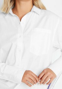 Woman wearing a white button-up shirt with a chest pocket, hands gently clasped in front, against a plain white background.