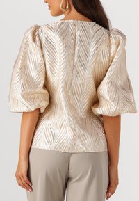Blouse met pofmouwen in lichtgoud met een gestructureerd, golvend patroon; heeft een ronde halslijn en een gladde achternaad. Gecombineerd met beige broek.