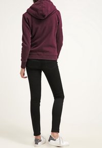 Sudadera con capucha color marrón granate de textura suave, combinada con vaqueros negros ajustados y zapatos blancos que tienen un detalle estampado en gris.