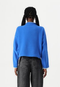 Modrý fleecový svetr s krátkým střihem, dlouhými rukávy a kulatým výstřihem. Ozdobený světle modrými okraji a kombinovaný s tmavými džínami.