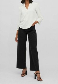 Blouse blanche texturée avec un col en V, associée à un pantalon large noir taille haute et des talons hauts noirs à lanières.