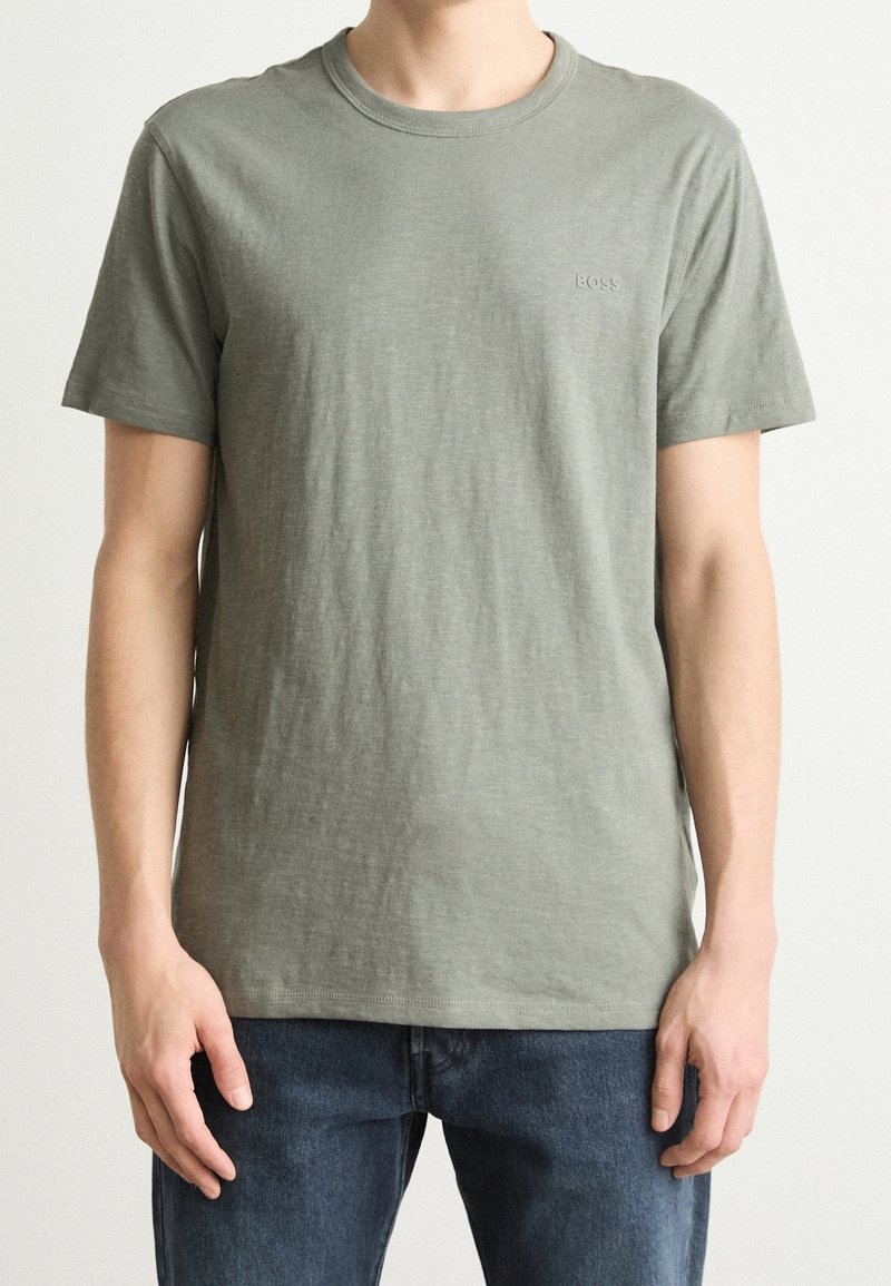 Homme portant un t-shirt uni vert olive à manches courtes et col rond et un jean bleu foncé, debout les bras détendus le long du corps.