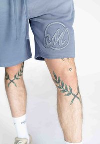 Grå atletiske shorts med broderet "M" logo på venstre ben, bæret med hvide sokker og stylet med grønne bladvævninger på benene.