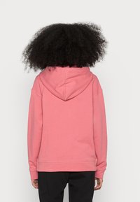 Osoba s kudrnatými vlasy v jednoduché růžové mikině a černých kalhotách, stojí otočená zády proti jednoduchému světlému pozadí.