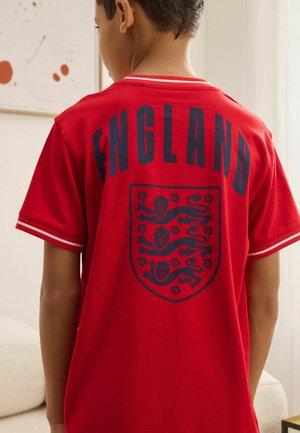 Gyerek piros rövid ujjú pólót visel, melyen az "ENGLAND" felirat és három oroszlán embléma látható a hátoldalon, bent áll fehér bútorok mellett.