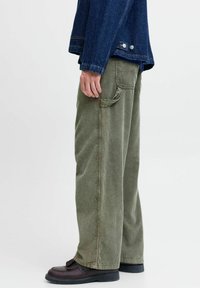 Grønne kordfløyelbukser med vide ben og sidelommer, kombinert med mørke sko. Materialets struktur er synlig med vertikale riller.