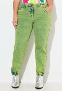 Pantalones jogger de denim verde con una textura desvaída, puños ajustados y diseño de cinco bolsillos. Combinados con botas de tobillo metálicas plateadas.