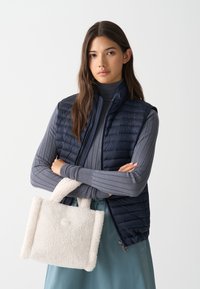 Tengeri kék puffervállú mellény steppelt textúrával, bordázott szürke pulóver és egy fehér műszőrme kézitáska kicsi fogantyúkkal és kerek logó részlettel.