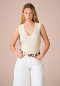 Top senza maniche color crema a coste con scollatura a V. Abbinato a pantaloni bianchi larghi e una cintura a strisce marrone e bianca.
