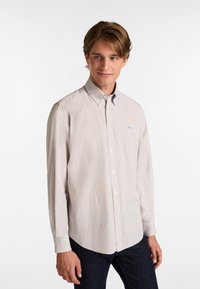 Camisa de botones de color beige claro con un sutil patrón a rayas, mangas largas y un pequeño logo azul en el lado izquierdo del pecho. Textura de tela suave.