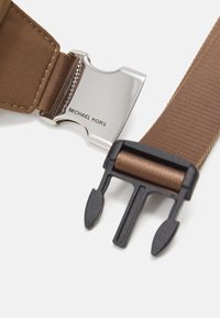 Brun stoffbelte med en sølvfarget metallspenne gravert med "MICHAEL KORS" og en svart plastlås med to klør.