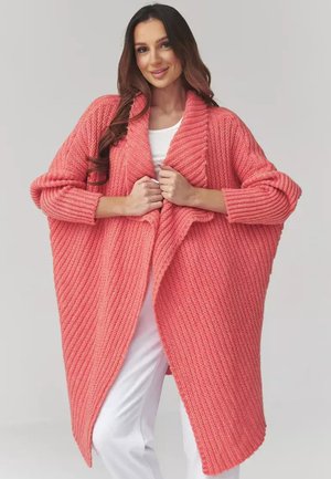 Kobieta w dużym różowym koralowym swetrze o fakturze dzianiny na biały strój, trzymająca przednie brzegi obiema dłońmi.