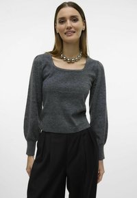Grauer Strickpullover mit langen, puffigen Ärmeln und einem quadratischen Ausschnitt, kombiniert mit hoch taillierten, gefalteten schwarzen Hose. Massiver silberner Halsreifen.