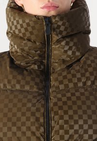 Brun dunjakke med ternet mønster, høj krave og lynlås foran. Materialet ser glat og skinnende ud med en polstret tekstur.
