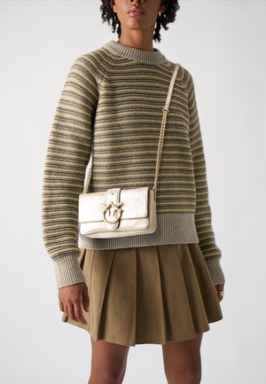 Borsa a tracolla metallica color oro con chiusura frontale, caratterizzata da una texture liscia e una tracolla a catena. Indossata insieme a un maglione a righe e una gonna plissettata.
