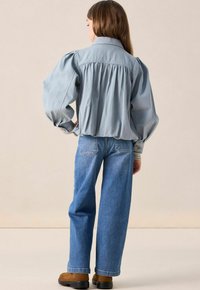 Jeune personne aux cheveux longs, portant une blouse légère bleu clair, un jean bleu droit et des chaussures marron, se tenant les mains sur les hanches, de dos.