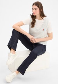 Jongere vrouw met lang bruin haar zit op een witte blok, gekleed in een witte T-shirt met korte mouwen, een grijs mouwloos vest, donkere jeans en witte sneakers.