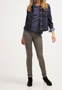 Kobieta w ciemnej dżinsowej kurtce, granatowej bluzce w kropki, oliwkowych wąskich spodniach i czarnych botkach za kostkę, stoi w odprężonej pozycji.