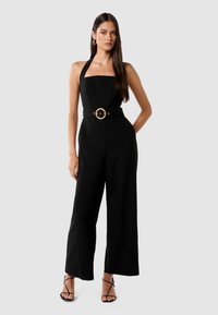 Svart jumpsuit med firkantet utringning, halterneck-stropper, vid ben-design og innsvingt midje med et stort, rundt gullbelte-spenne.
