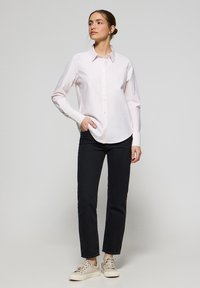 Camicia button-up rosa chiaro con colletto, maniche lunghe e sottili righe, abbinata a jeans slim-fit neri e scarpe da ginnastica beige.