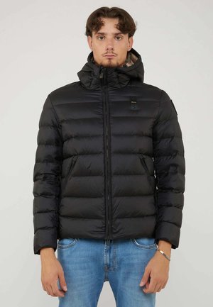 Junger Mann mit Blick nach vorne, trägt eine schwarze Steppjacke mit Kapuze und blaue Jeans, steht vor einem schlichten hellen Hintergrund.