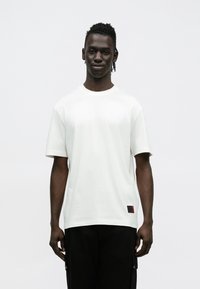 Άσπρο βαμβακερό T-shirt με κο�ντά μανίκια, στρογγυλή λαιμόκοψη και ένα μικρό κόκκινο λογότυπο στην κάτω αριστερή πλευρά. Φοριέται με σκούρο παντελόνι.