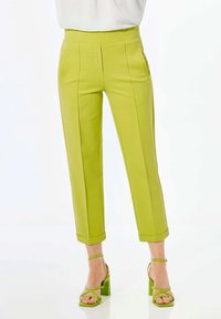 Grüne Cropped-Hosen mit hohem Bund, seitlichen Taschen und hochgekrempelten Säumen. Kombiniert mit grünen High-Heels. Glatte Textur.