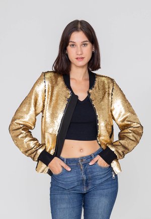 Giovane donna con capelli scuri indossa una giacca di paillettes dorate lucida sopra un crop top nero e jeans blu, in piedi con le mani in tasca.