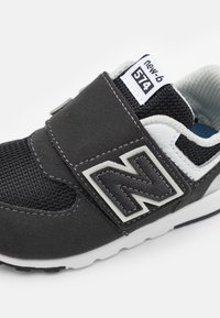 Crna atletska obuća s gornjim dijelom od sive nubuk kože, mrežastim umetcima, čičak remenom i bijelim detaljima. Sadrži logo New Balance u sivoj boji.